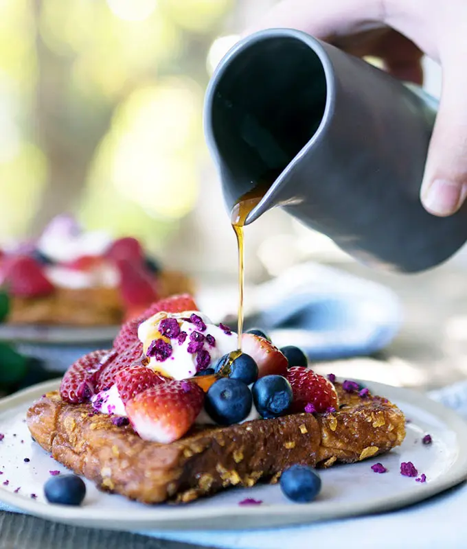 maple syrup being poured from a stone blue jug over cornflake crumbed french toast topped with berries and yoghurt