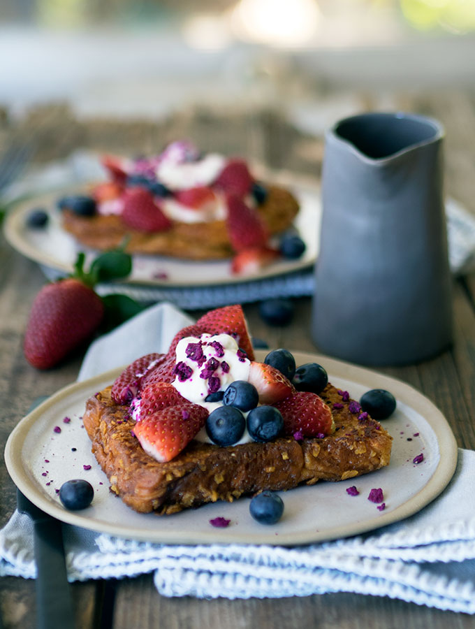 french toast toped with berries and yoghurt on a plate on a wooden table with jug of maple syrup