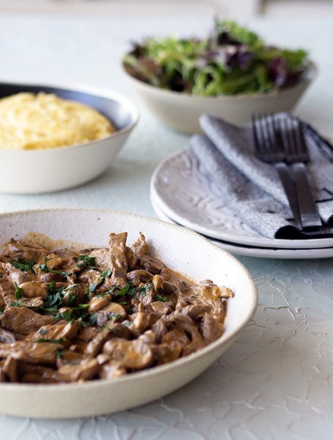 boeuf stroganoff in a ceramic bowl on table with plates, bowl of polenta and bowl of salad in the background