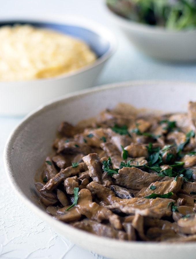 close up of beef stroganoff in a ceramic bowl on table, bowl of polenta in the background out of focus