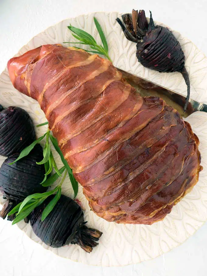 prosciutto wrapped turkey breast with tarragon citrus butter just out of the oven on a platter with hasselback beetroot and sprits of tarragon
