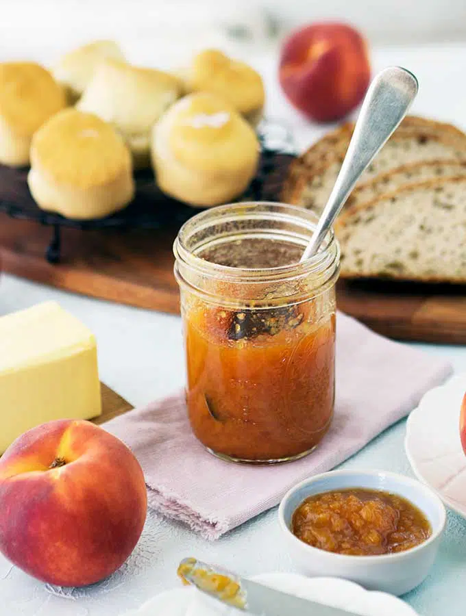jar of peach bourbon jam on a linen serviette surrounded by scones, bread and peaches