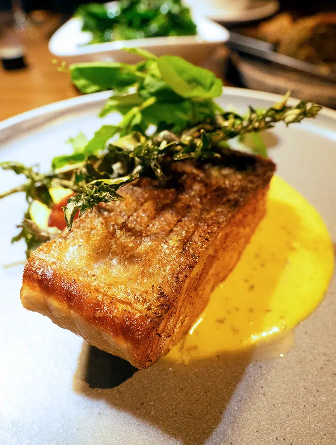 a piece of salmon with crispy skin sitting on hollandaise sauce with greens on top