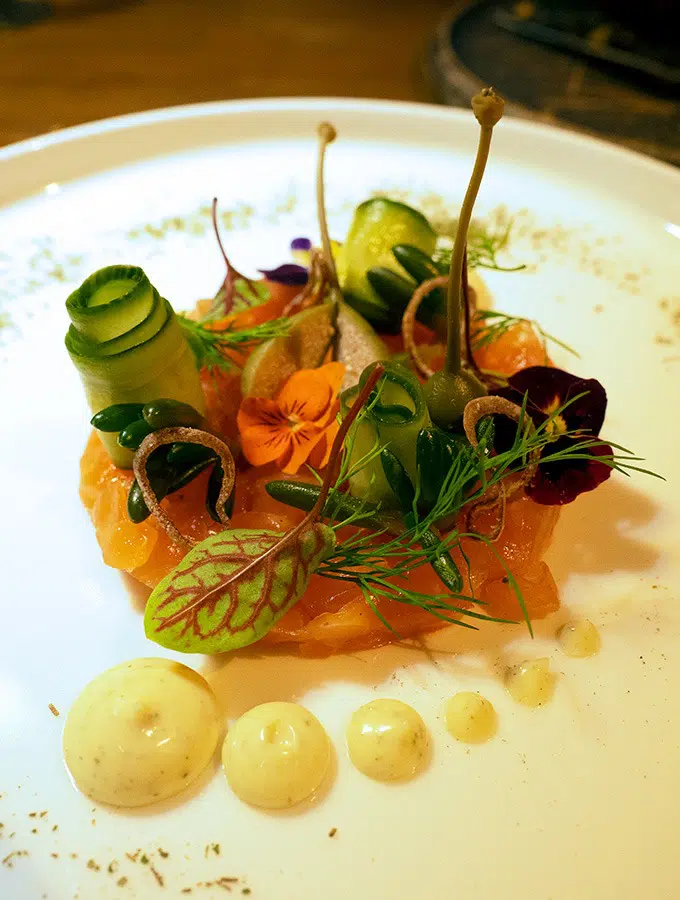 cured salmon on a plate with curls of cucumber, caperberries, flowers, micro herbs and dots of aioli at the front