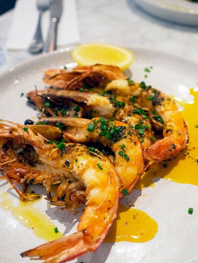 grilled large prawns on a plate covered with a sauce and a slice of lemon at the back