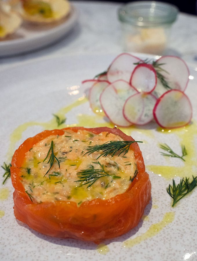 salmon rillett wrapped by smoked salmon with herb cream at the back that has been decorated with thin slices of radish.