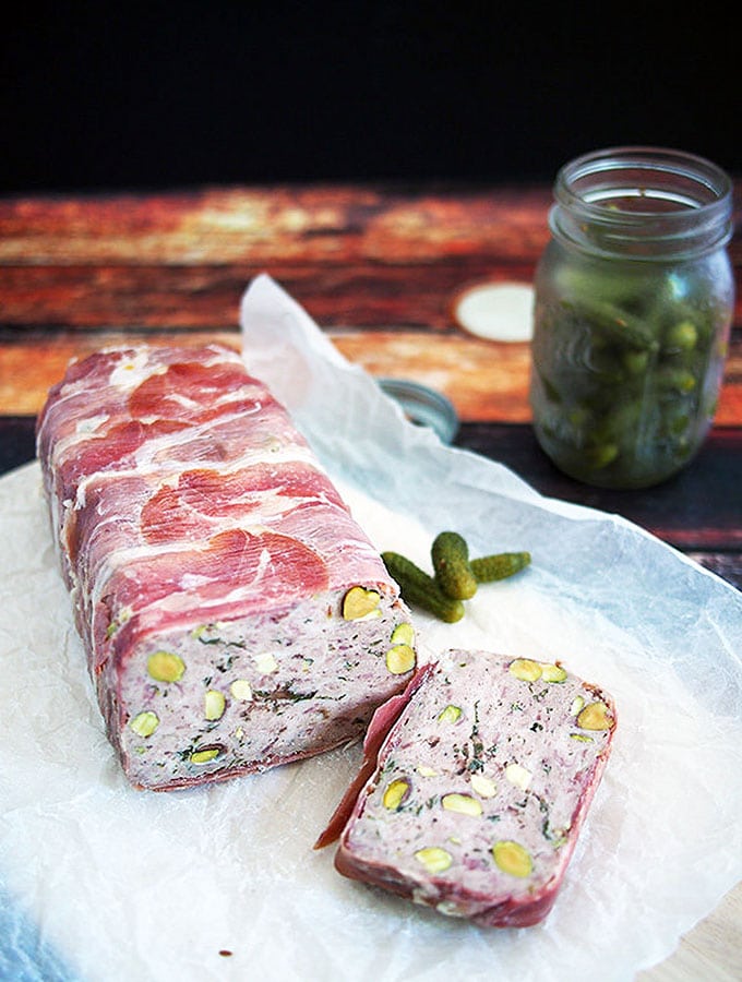 pork and veal terrine studded with pistachios sliced on paper with a jar of cornichons in the background and a couple next to the terrine