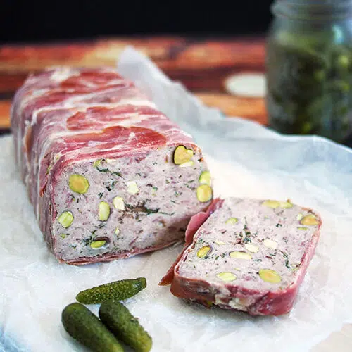 pork and veal terrine studded with pistachios sliced on paper with a jar of cornichons in the background and a couple next to the terrine