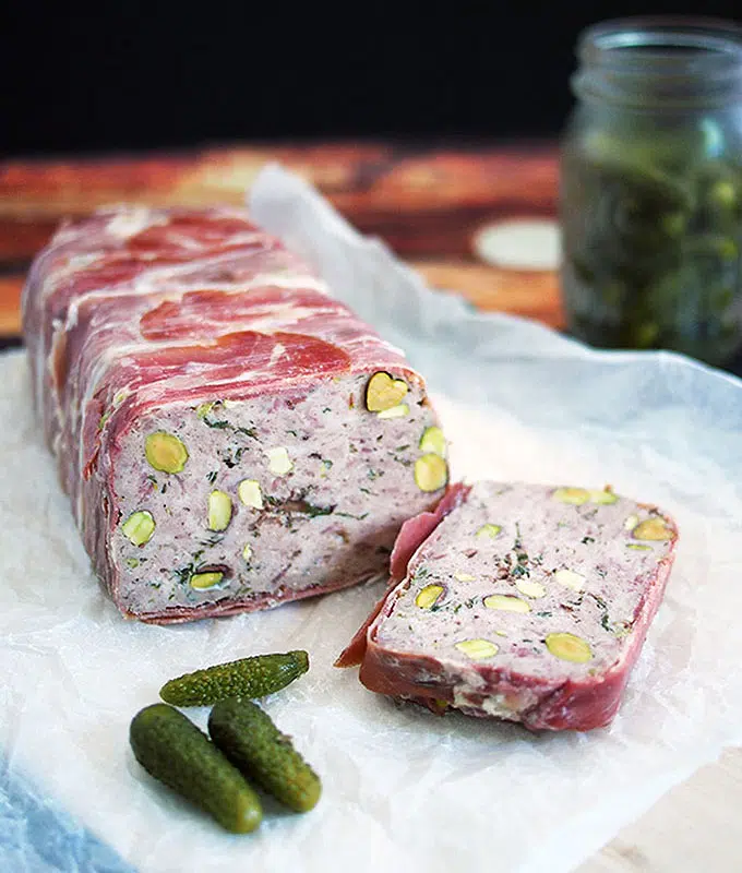 pork and veal terrine studded with pistachios sliced on paper with a jar of cornichons in the background and a couple next to the terrine