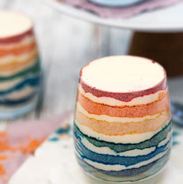 Thee glasses of rainbow coloured dessert with layers of coloured crushed biscuit and cream.