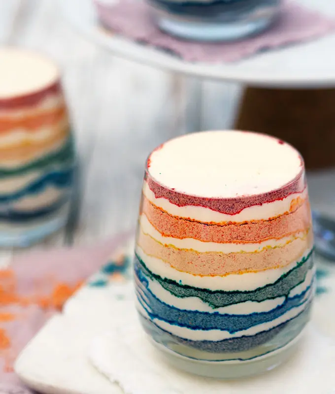Thee glasses of rainbow coloured dessert with layers of coloured crushed biscuit and cream.