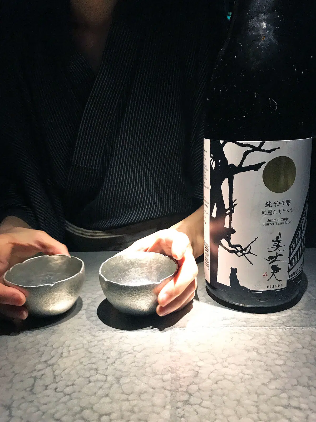 sake cups being presented by a bar tender on a tin counter next to a sake bottle