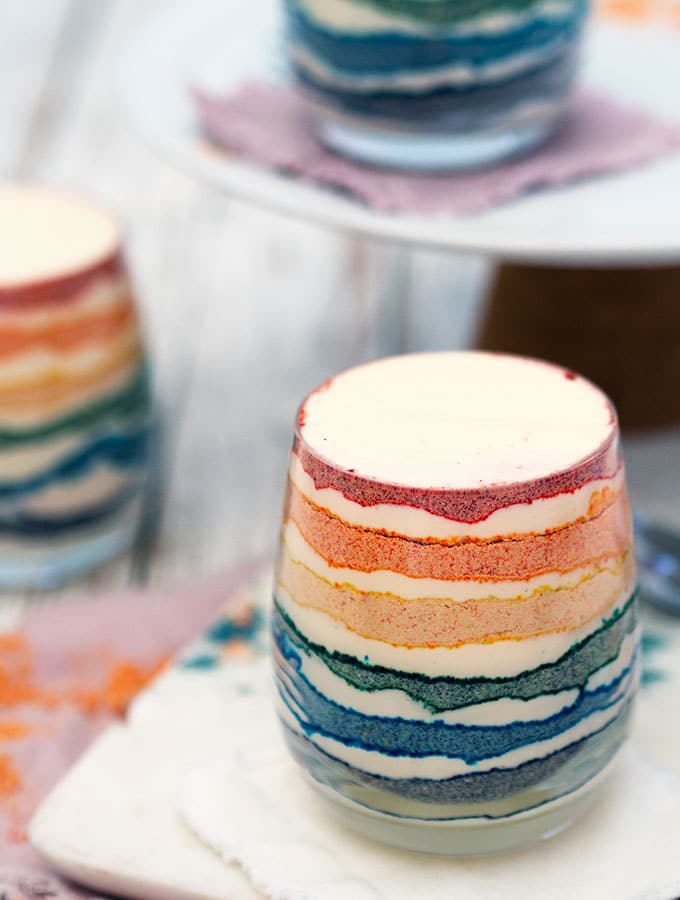 Close up of one glass of rainbow coloured dessert with layers of coloured crushed biscuit and cream.