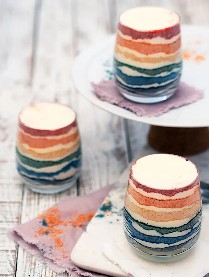 Thee glasses of rainbow coloured dessert with layers of coloured crushed biscuit and cream.