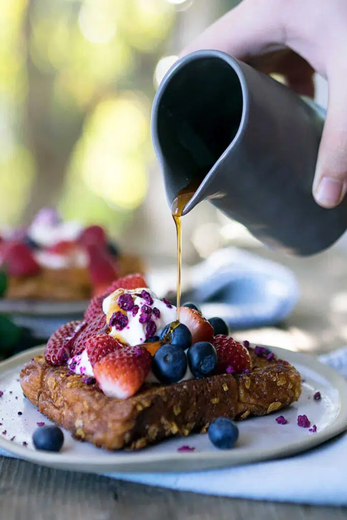 A person pouring maple syrup over french toast toped with berries and yoghurt on a plate