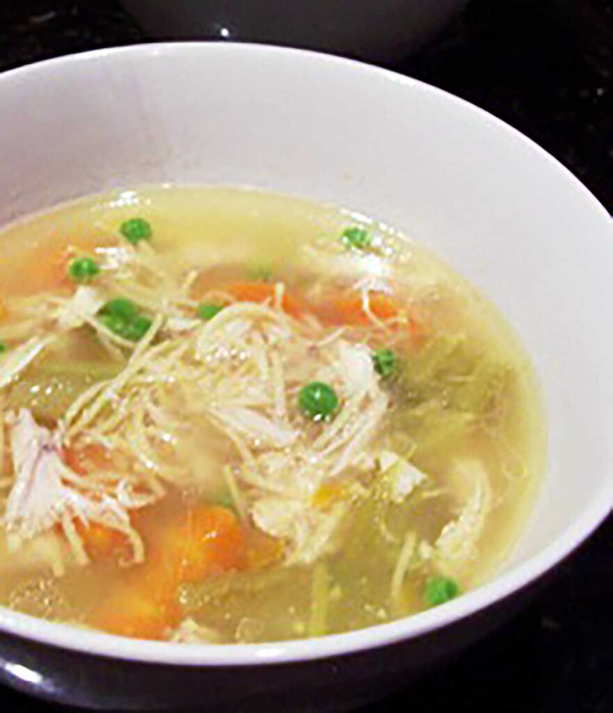 white bowl of chicken soup with thin noodles, carrots, celery and peas
