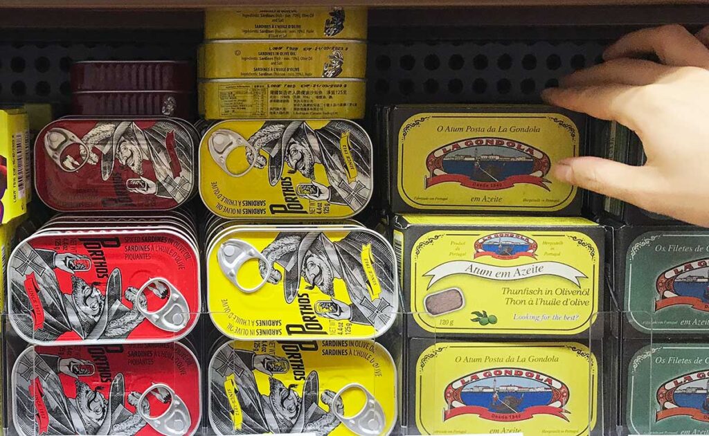 various colourful tins of sardines on a supermarket shelf with hand reaching in to grab one