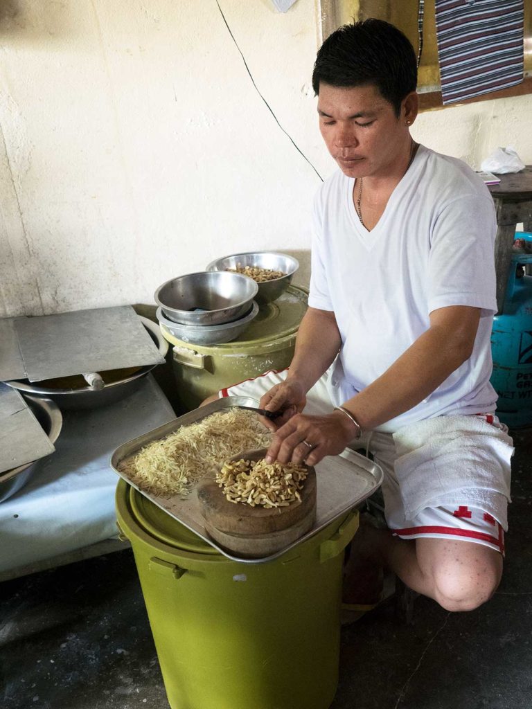 man sitting with a green bin between his legs, slicing cashews very finely with a knife