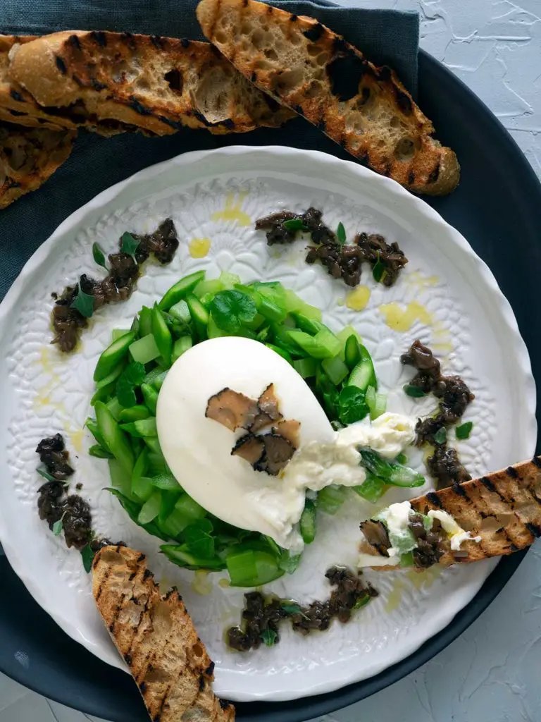 burrata on a bed of asparagus and celery, topped with truffle carpaccio, salsa tartufata dolloped around the outside on a white plate. grilled bread being used to eat it