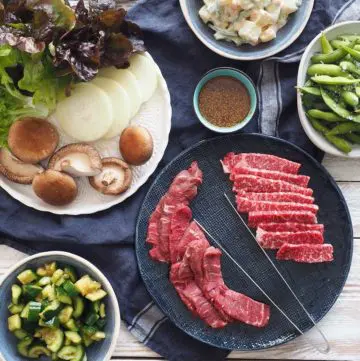 Ingredients for Yakiniku, beef slices on a plate, crushed cucmbers in a bowl, slices of onion, whole shitake mushrooms and lettuce on a plate, sauce in a bowl, cooked edamame in a bowl and Japanese potato salad in a bowl