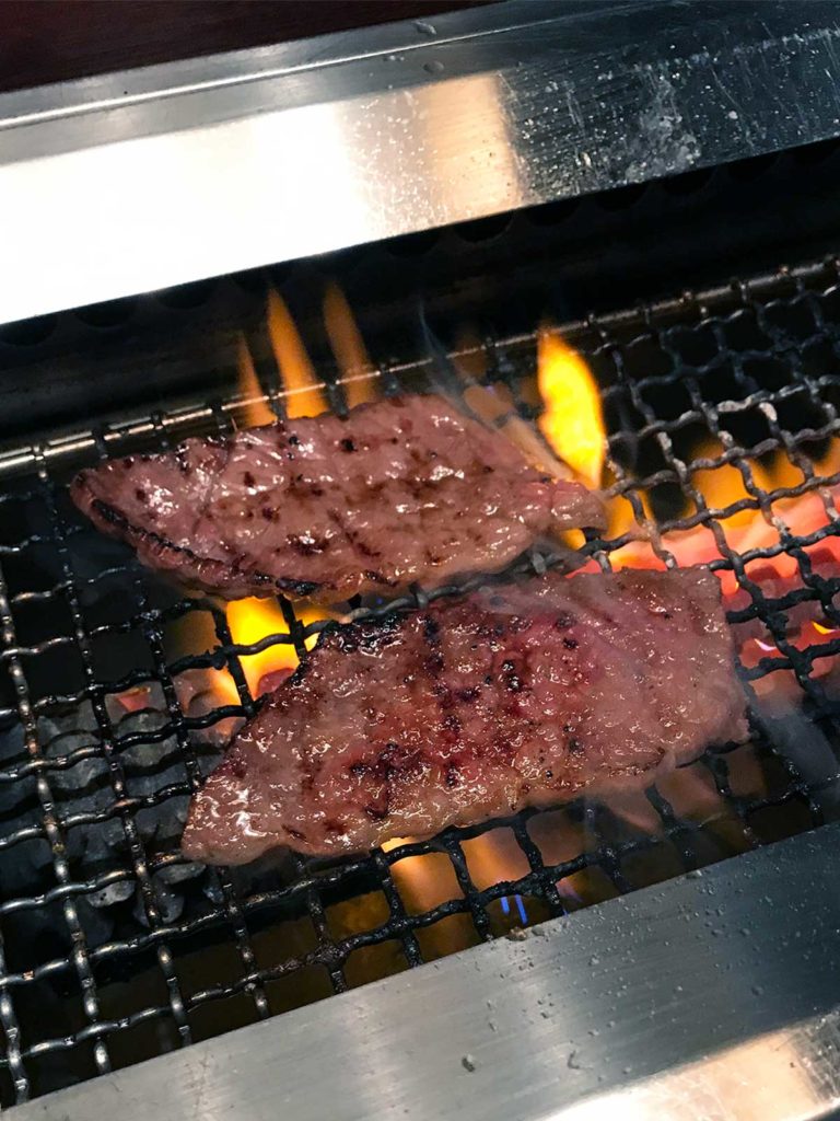 two pieces of thinly sliced steak being cooked on a gas bbq