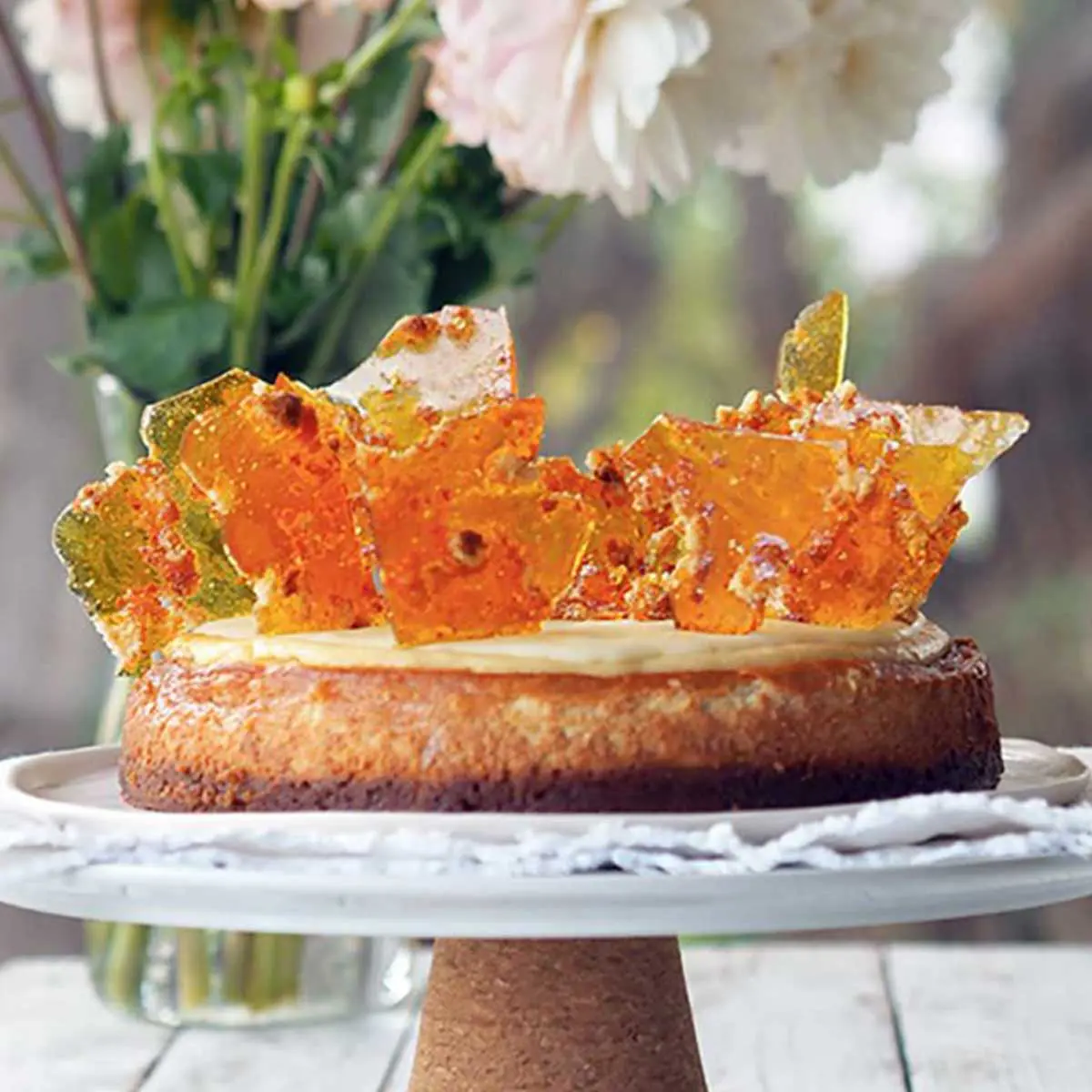 Cheesecake topped with shards of toffee, place on top of a cake stand, flowers and greenery adorn the background