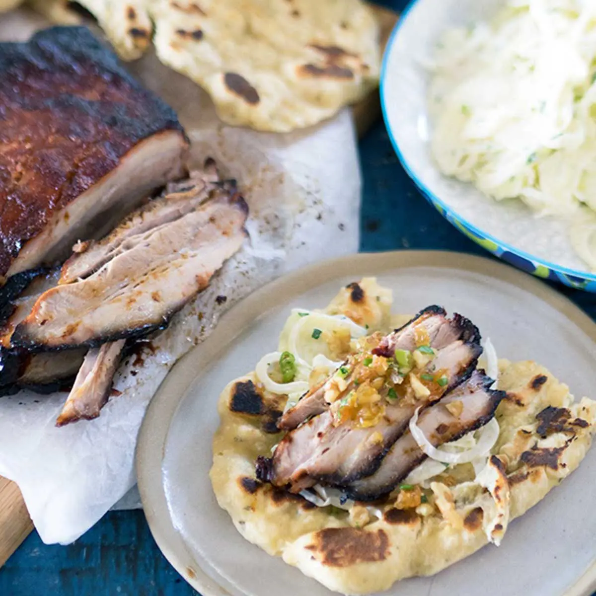 Slices of pork belly with slaw on top of flatbread topped with walnut crumble, pork belly and extra flatbread in the background