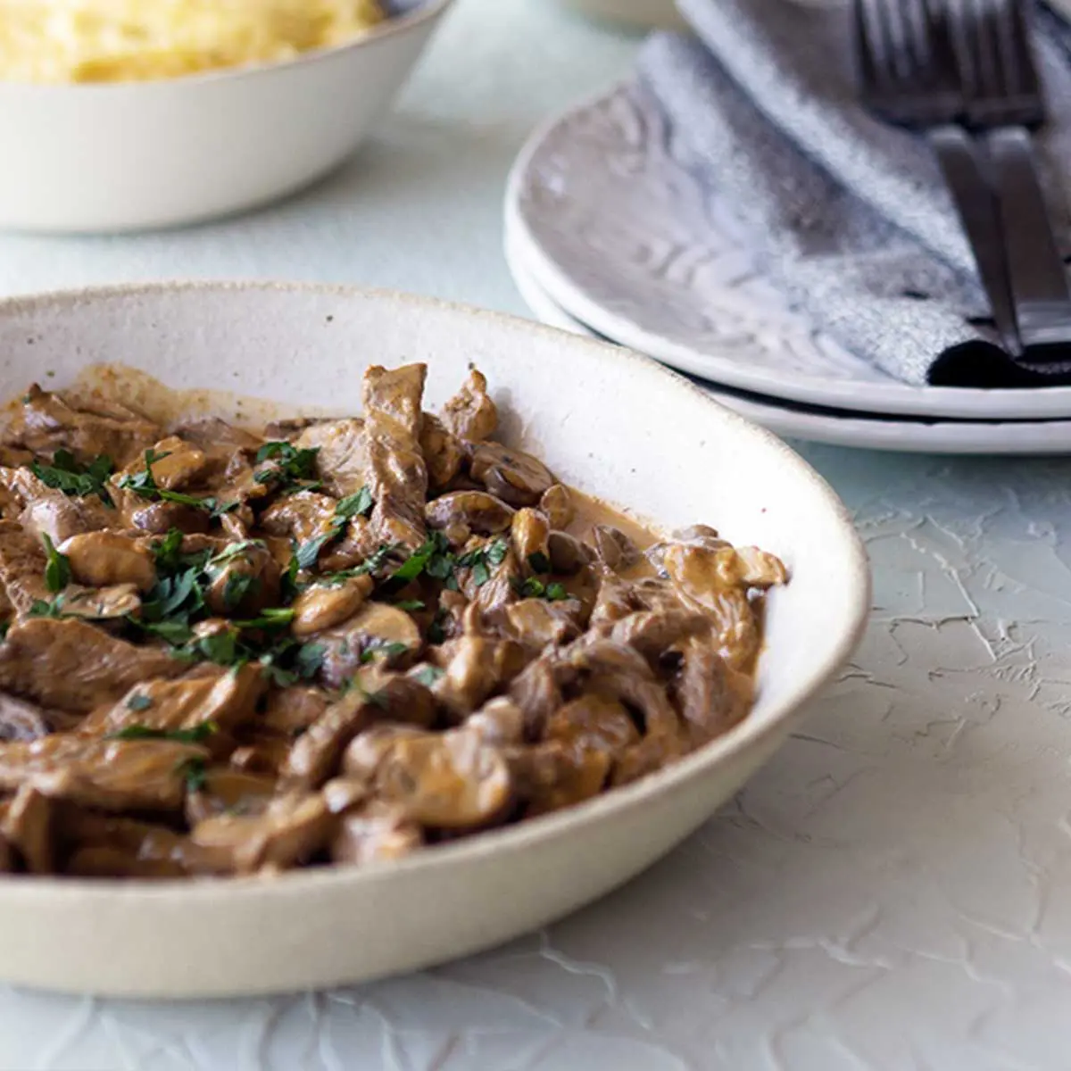 a stone bowl of beef and mushroom Stroganoff or stew, background is a textured white surface and stacked plates with forks