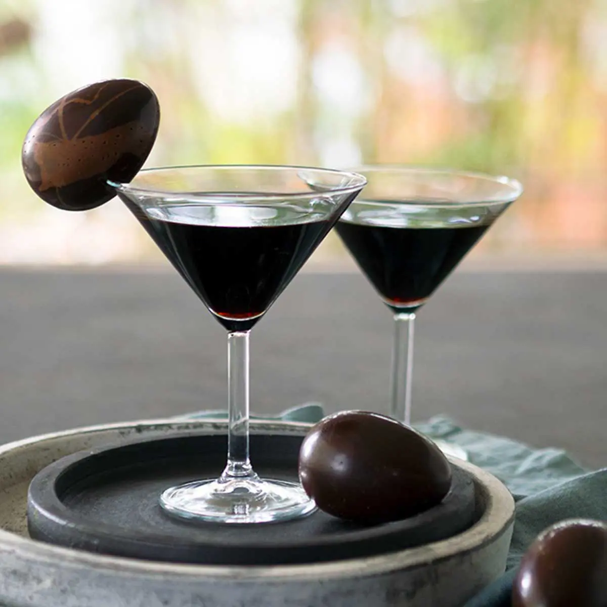 Dark almost black cocktail in a martini glass garnished with a dark chocolate egg on the rim
