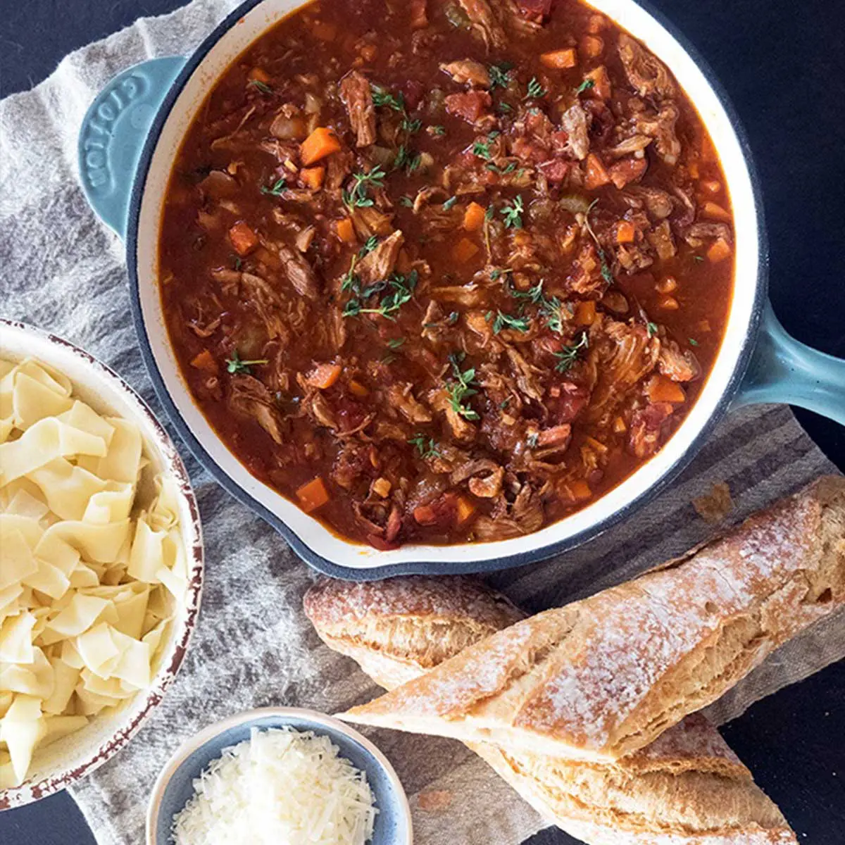 A dark duck ragu in an iron pan place atop a tea towel, crusty bread, pasta and shredded cheese accompany it.