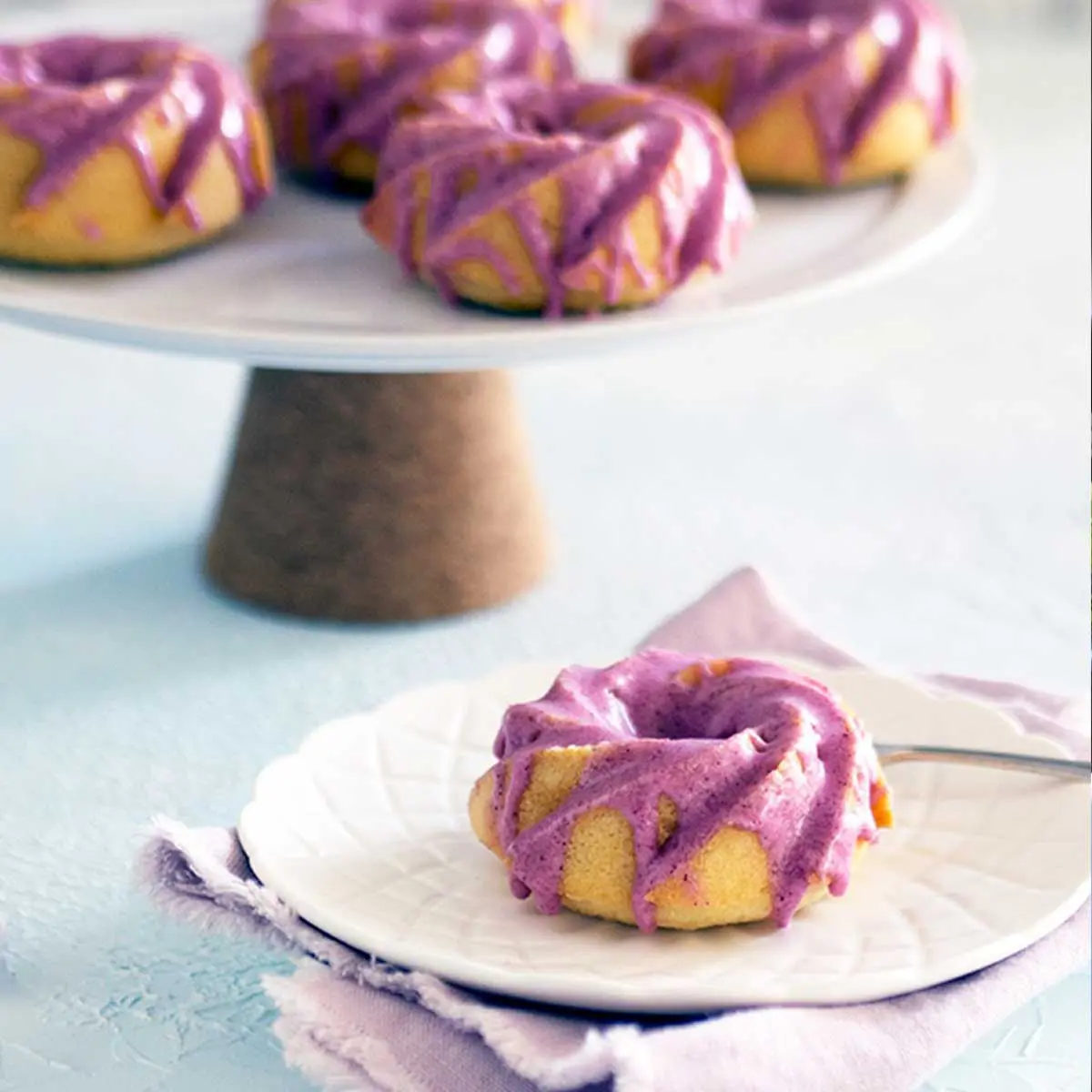 Small cake drizzled with icing on a white plate, additional cakes on a cork based cake stand in the background