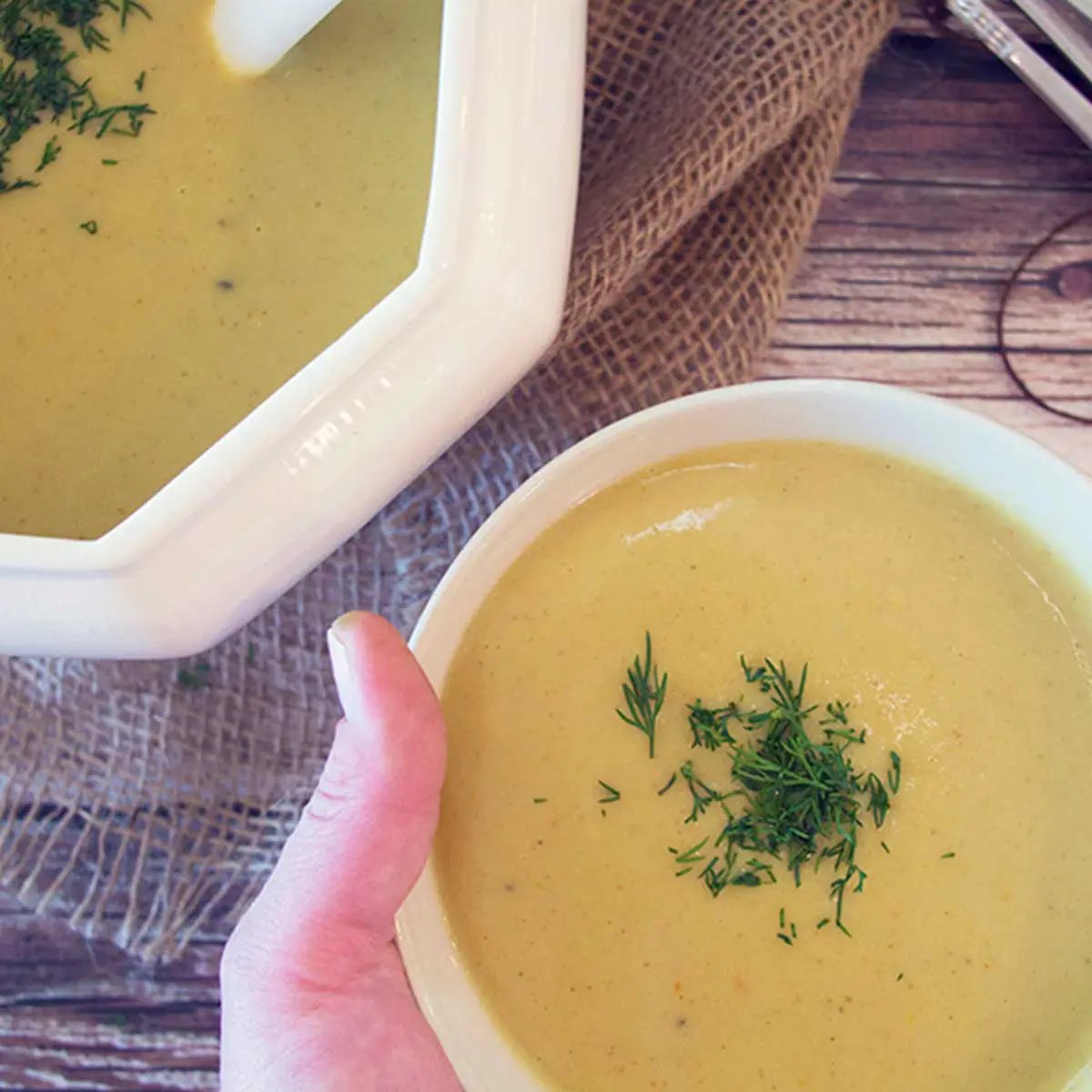 A white bowl holding a yellow-orange serving of soup garnished with chives, burlap and wood in the background