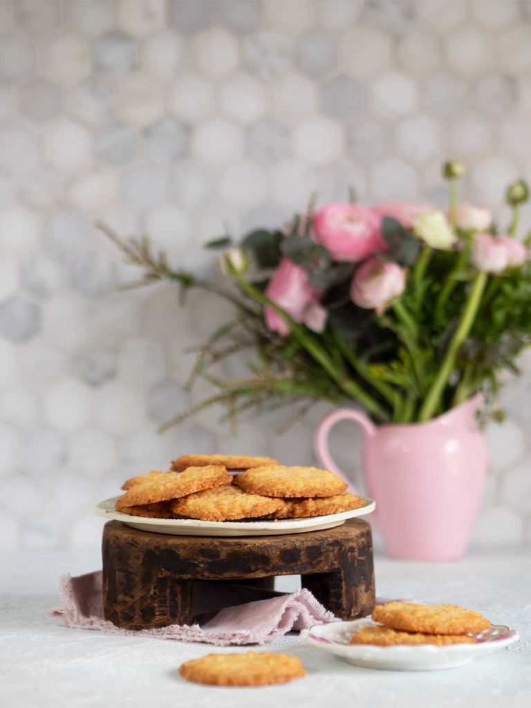 coconut cookies on a plate on a wooden stand, some more cookies in front and vase with pink flowers behind