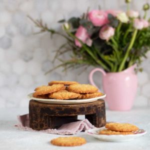 coconut cookies on a plate on a wooden stand, some more cookies in front and vase with pink flowers behind