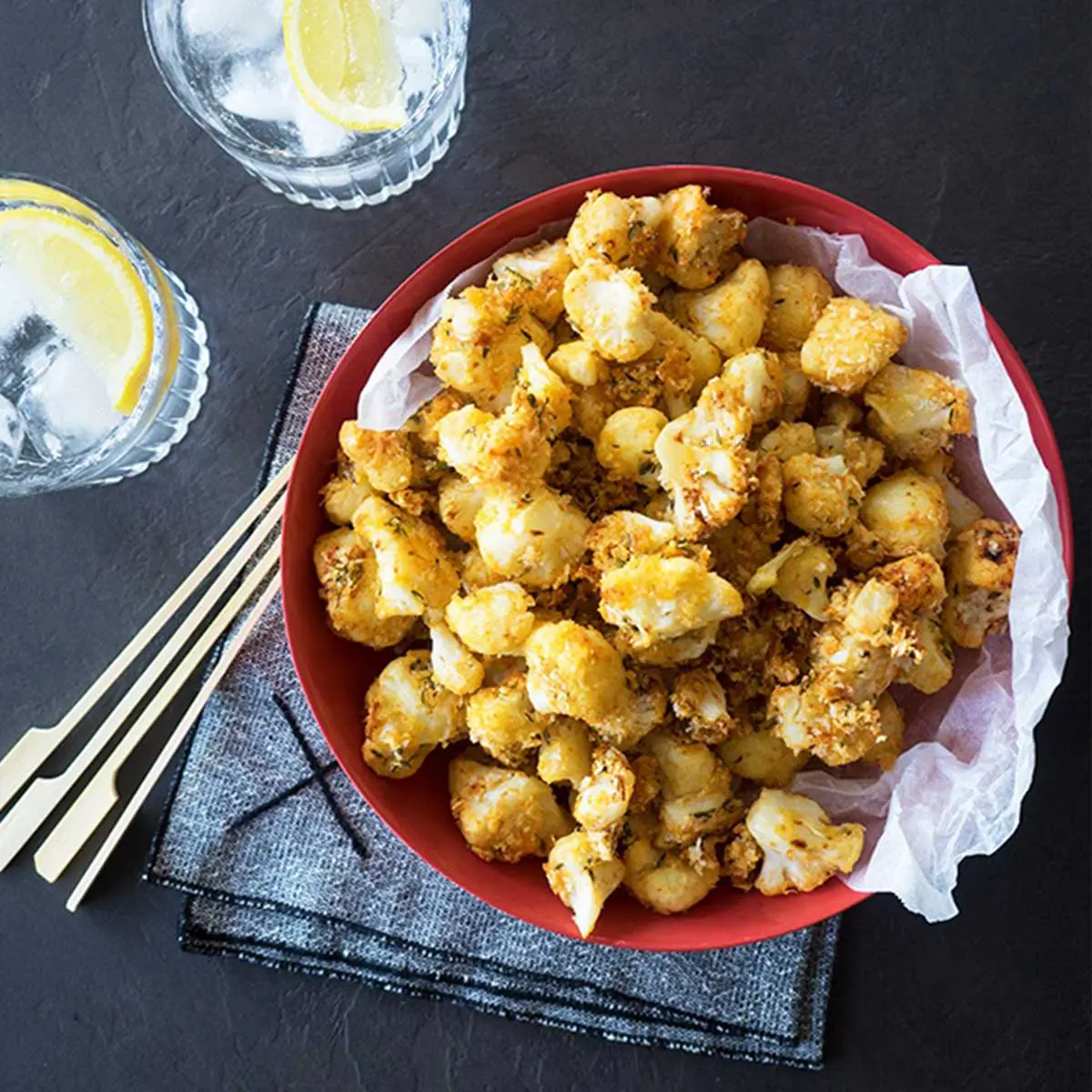 A red bowl filled with crunchy oven baked crumbed Cauliflower florets, 2 glasses of water rest on a dark grey textured background