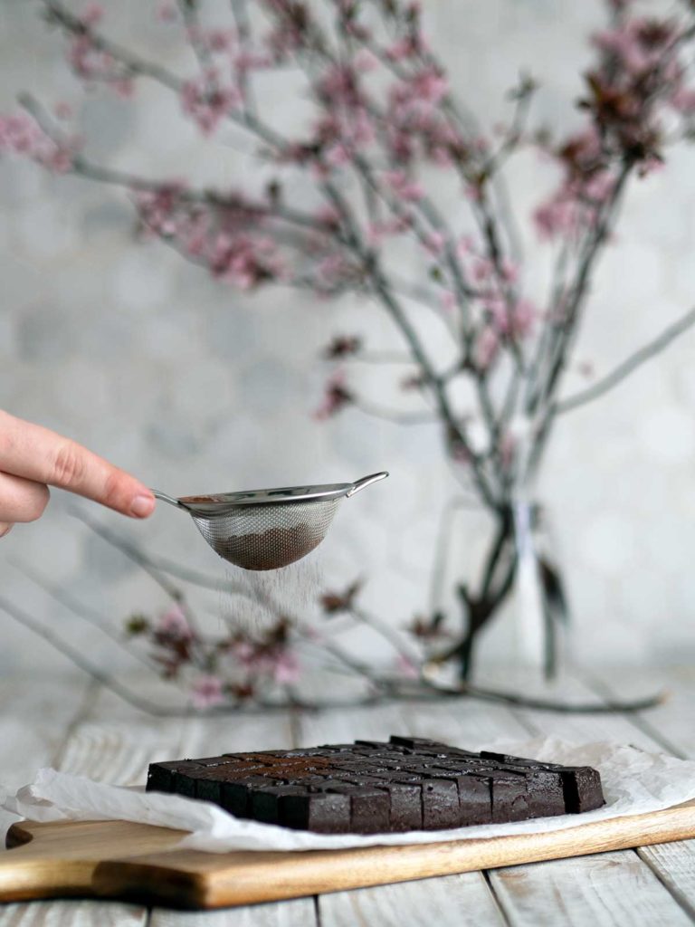 cocoa powder being sifted over the top of cut nama chocolate with cherry blossoms in the background