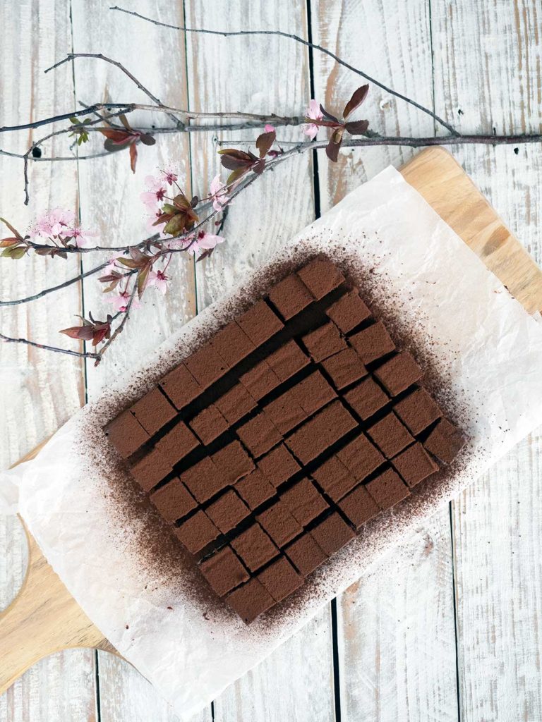 nama chocolate cut into squares dusted with cocoa powder on some paper on a wooden board with cherry blossoms in the top left hand corner