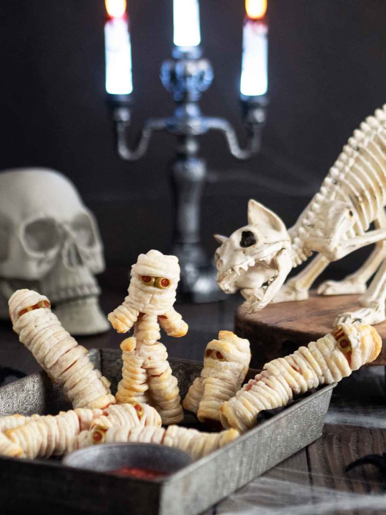 mummy hot dogs in a metal containers with a skeleton cat and skull in the back ground