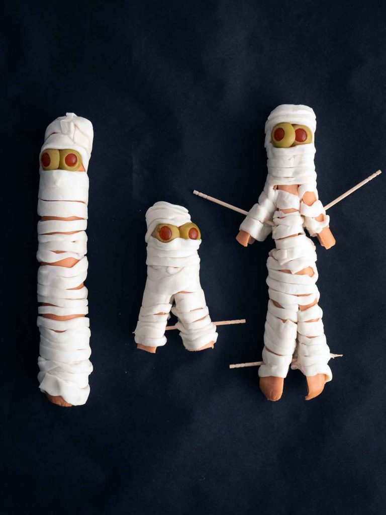 hot dogs wrapped up like mummies on black paper with toothpicks securing arms and legs