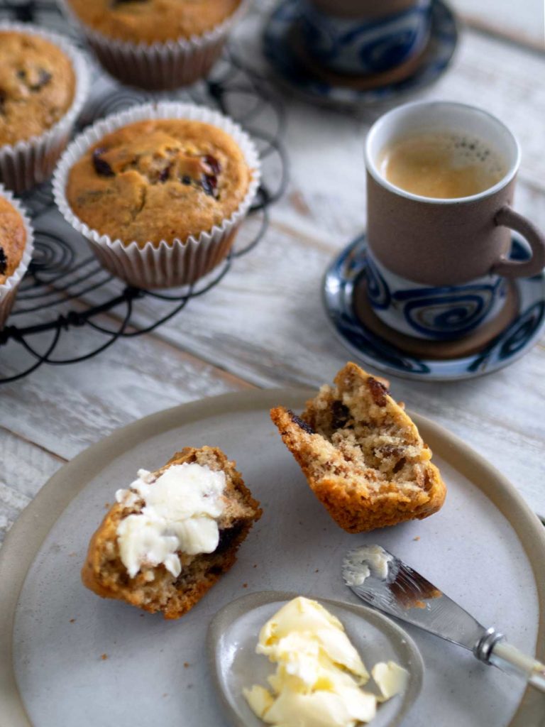 muffin split open on a plate spread with butter, cup of coffee and tray with muffins in the background
