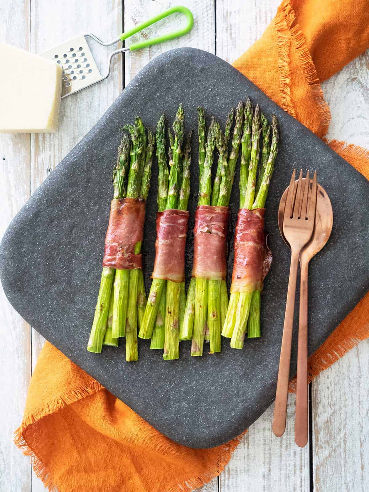 Undressed asparagus bundles on a black plate with serving spoons.