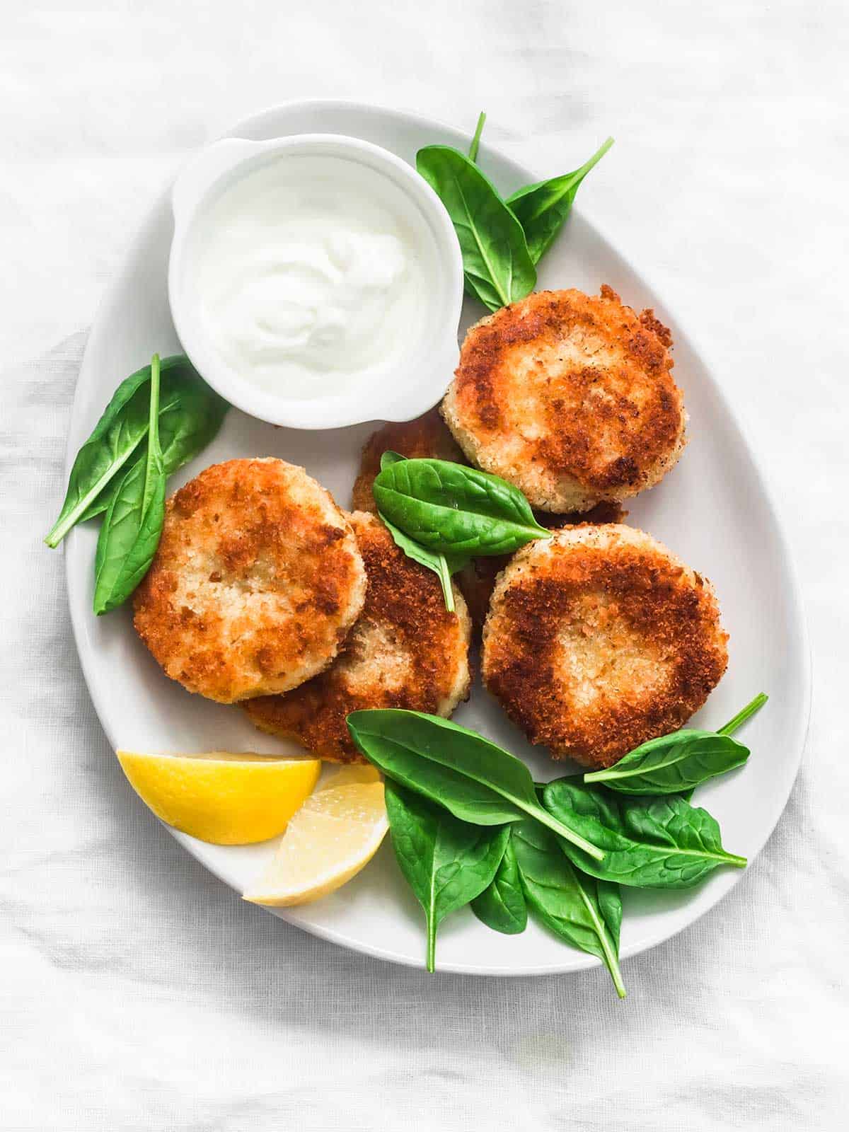 Salmon patties on a white plate with baby spinach, lemon wedges and white sauce in a bowl.
