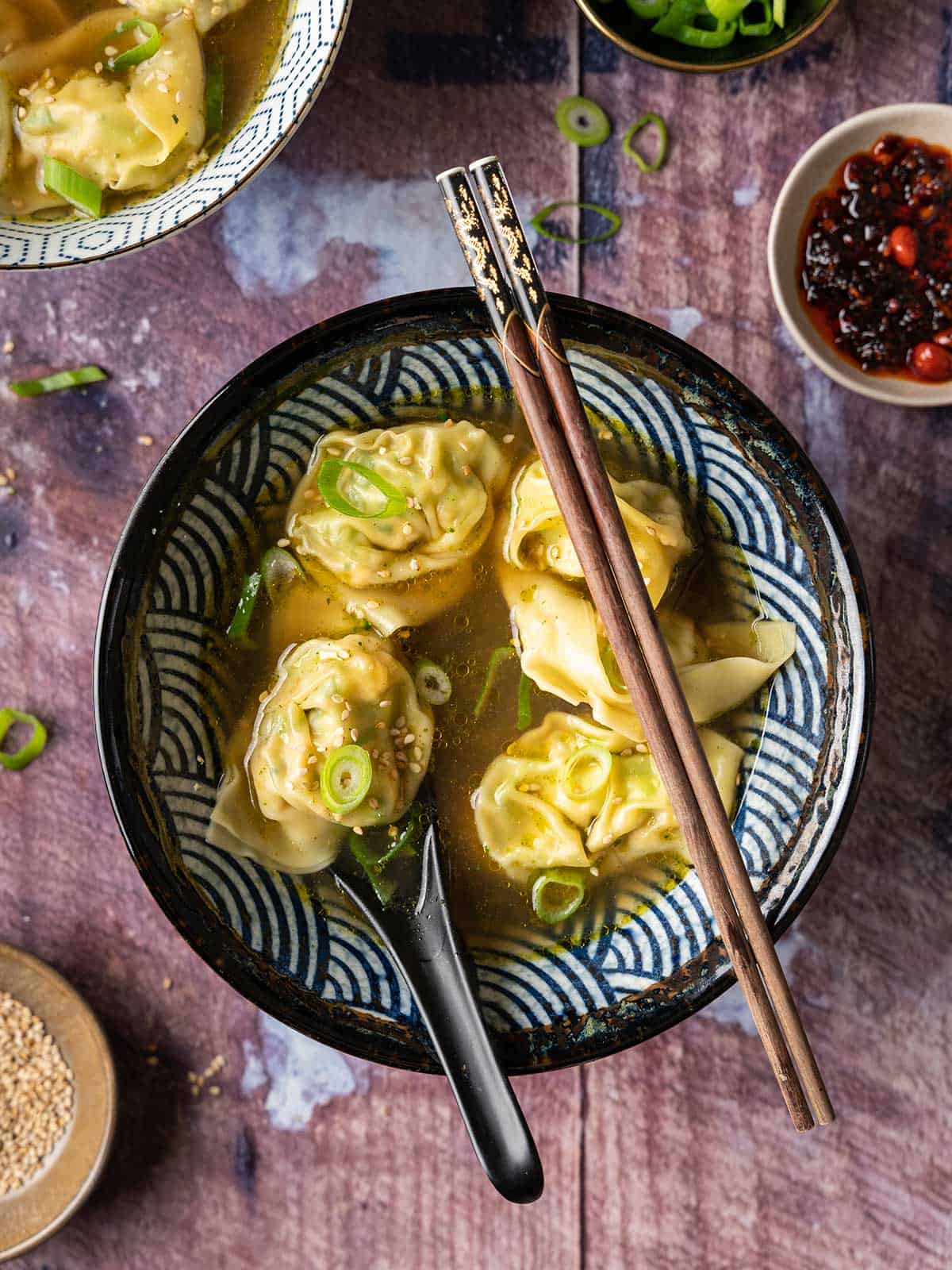 Wontons and soup in a black and white bowl with chopsticks and a spoon.