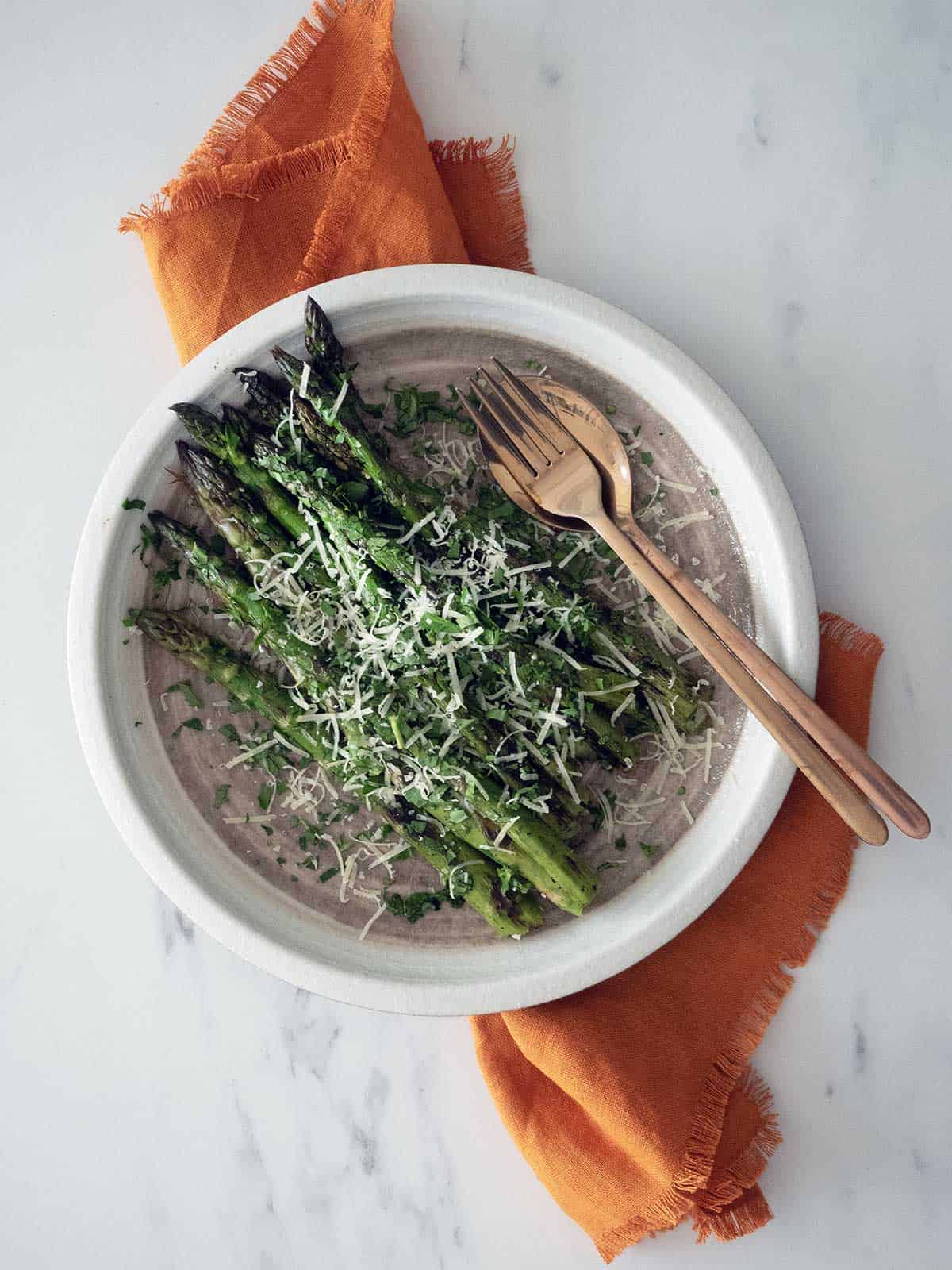 Grilled asparagus on a white plate with serving fork and spoon.
